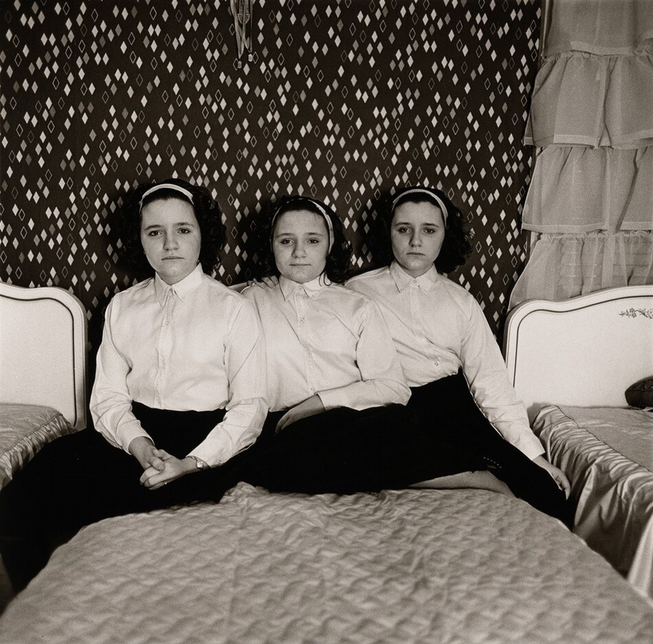 In this black-and-white photograph, three identical young women are seated side by side on a bed in a bedroom. They are positioned closely together, facing the camera. Each has an oval face, pale skin, and wavy dark hair pulled back with a white headband, and they are all wearing white shirts and dark skirts. The woman on the left has her hands clasped in her lap. The room has patterned wallpaper with diamond-like designs on a dark background, and on either side of the central bed, partial views of bed headboards and ruffled curtains are visible.