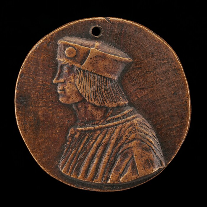 The sculpture is a round medallion featuring a detailed side profile of a man's head. The subject is depicted wearing a brimmed hat that covers his hair, which falls in a fringed style to just above his shoulders. He is also wearing a pleated garment that can be seen at the shoulders and upper torso. The material of the sculpture appears to be bronze with a warm brown patina. The medallion has a circular hole near the top. The features of the face, the texture of the hair, and the folds of the garment are intricately rendered, creating a clear representation of the subject in profile against the flat background of the medallion.