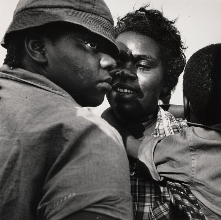 The image shows three individuals in a close-up shot. On the left, a person is seen from the side, wearing a hat that casts a shadow over their eyes. Their facial expression is neutral. The central figure, a woman, has partially closed eyes and smiling lips. Her hair is styled in short, curly locks, and she wears a checkered shirt. A child's arm is around her shoulder, with the child's head partially visible to the right. The background is plain and light, emphasizing the foreground figures.