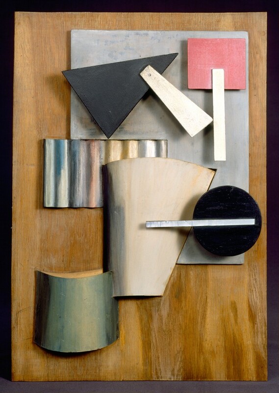 A collection of geometric shapes, many painted a single color, are arranged on a background painted to look like wood grain in this abstract vertical composition. A silvery gray, upside down, wide L shape fills most of the upper half to our right of center. On it, a rose-pink square is attached to an ivory-white rectangle suggestive of a handle, and a black triangle with a similar handle sits on the L form to our left. A rectangular piece of corrugated silver metal is tucked into the corner of the L shape. A black circle, a white conical form, and a curving, sage-green piece create a row from right to left down into the lower left corner of the composition.