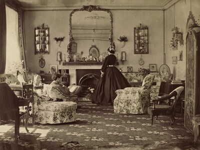 “Interior of Room”. We look toward the far end of a large, well-appointed room at a person reading in an armchair and a woman wearing a long dress standing in front of a fireplace in this horizontal photograph.