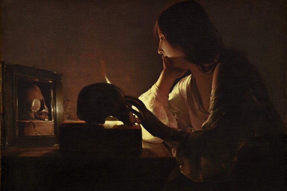 A young woman with pale skin sits at a table in a darkened room in this vertical painting. Long chestnut-brown hair drapes over her shoulder and her deep, cream-colored, long-sleeved garment is open at her neck. She rests her chin in her right hand, farther from us, as her left reaches for a skull placed on a thick book on the table in front of her. The scene is lit by a single candle mostly out of sight behind the skull. Shown in profile, she looks into a small mirror next to the skull, which reflects that object and the book.