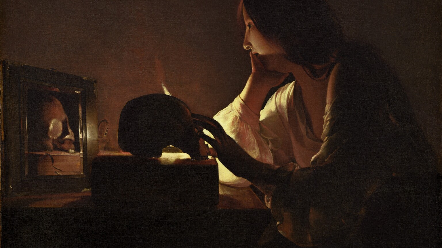 A young woman with pale skin sits at a table in a darkened room in this vertical painting. Long chestnut-brown hair drapes over her shoulder and her deep, cream-colored, long-sleeved garment is open at her neck. She rests her chin in her right hand, farther from us, as her left reaches for a skull placed on a thick book on the table in front of her. The scene is lit by a single candle mostly out of sight behind the skull. Shown in profile, she looks into a small mirror next to the skull, which reflects that object and the book.