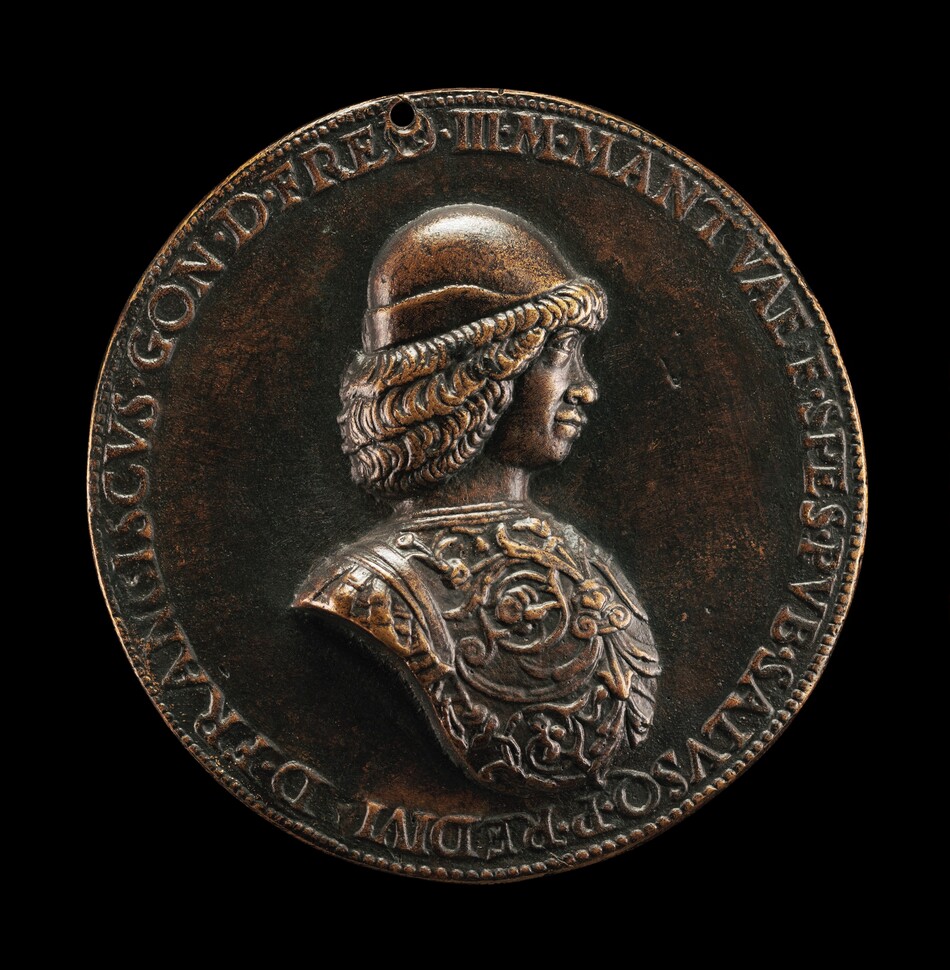 The sculpture is a relief depicting a male figure in profile, with curly hair flowing beneath a helmet. He wears detailed armor and the piece is circular in shape. It is crafted from bronze, with a dark metallic hue and subtle highlights. The rim is inscribed with text, and there is a small hole at the top. The sculpture has a glossy and smooth finish.