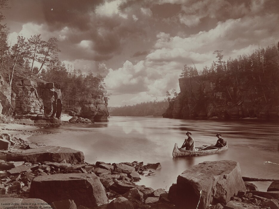 Two men in a canoe row toward us and a bank of rocks and boulders lining the bottom edge of this black and white, nearly square photograph. Sheer cliffs and trees rise from the banks to each side. The cliffs narrow like an almost closed letter C a short distance from us and then open onto wider tree-lined banks beyond. Puffy clouds skim across the sky above.