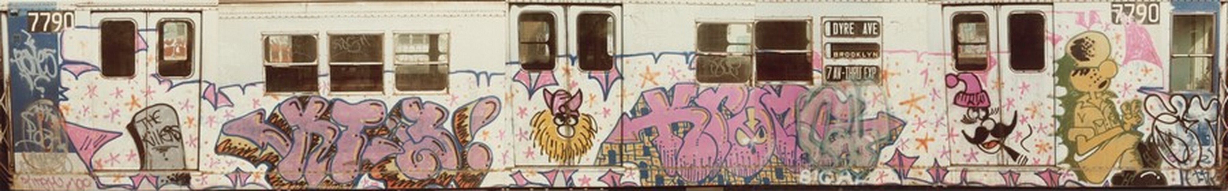 This image shows a subway train covered in vibrant graffiti. The train, numbered 7790, displays colorful street art featuring bold letters, cartoon characters, and abstract shapes in shades of purple, pink, and blue. The background is white with pink star-like elements. The windows and doors show signs of wear and additional graffiti tags. Characters on the train include one wearing a military-style helmet and holding a flower, and another with exaggerated features wearing a pointy hat. The scene conveys urban graffiti's raw artistic expression.