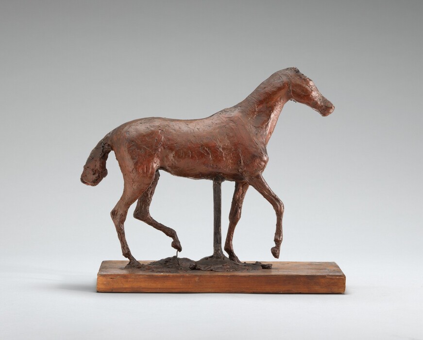 The sculpture depicts a horse in a pose suggesting movement. The figure is slightly abstract with a textured surface. The horse is primarily brown in color, resembling a bronze patina. It is mounted on a wooden base. The horse is presented in profile, displaying a slender body, elongated legs, arched neck, and detailed mane and tail. The piece has an unfinished, tactile quality.