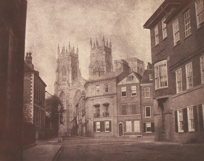 This sepia-toned photograph shows a street lined with buildings leading towards a distant cathedral.  The buildings closest to us appear to be made of brick and have white windows. The street leads off to the left, towards the grand cathedral in the background. The view is from street level, and the photograph has a grainy texture.