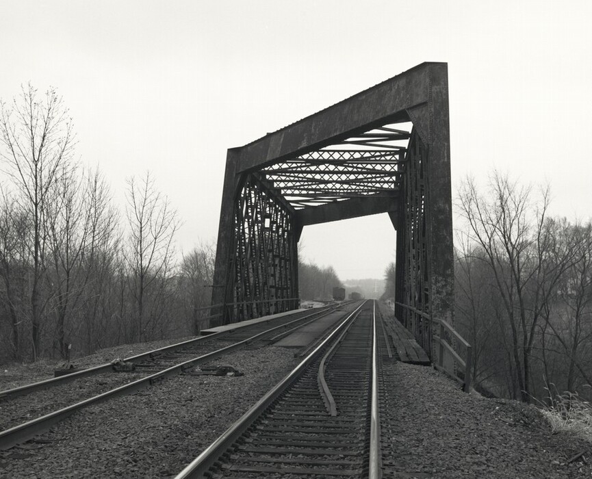 The image shows a railway track leading to a metal bridge. The tracks run straight ahead and disappear into the truss bridge, which dominates the scene with its industrial design. Beyond the bridge, the track continues into the distance until it meets the horizon, obscured by mist or fog. The sky above is overcast, offering a muted backdrop. The lack of color emphasizes the geometry of the bridge and tracks, while leafless trees line the railway, adding texture and contrast to the scene.