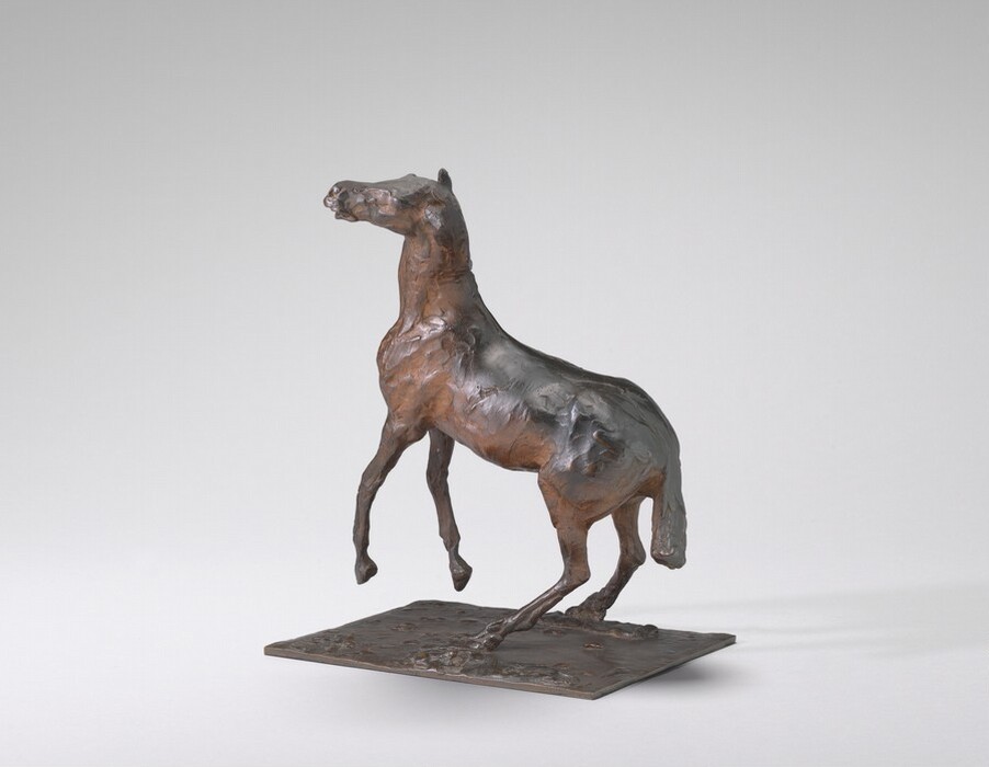 The sculpture depicts a horse in a raised stance on its hind legs, crafted from metallic material, possibly bronze. The color is warm brown with darker accents highlighting muscle texture. It is mounted on a flat base, with one hoof at the edge, suggesting motion. The detailed but slightly abstract form offers a lifelike representation of a horse's build and posture.