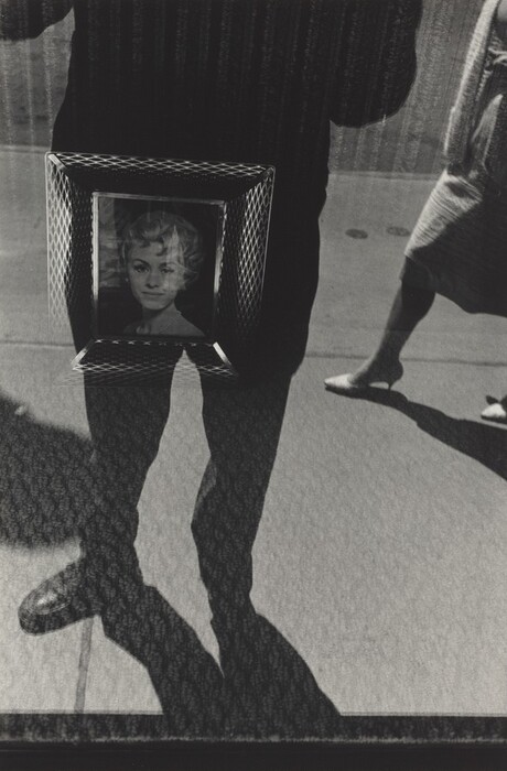 The image includes a complex arrangement of shapes and lines. It shows a distinct shadow of a person cast onto the sidewalk, intersecting with a framed portrait. This abstract composition blends together the real and the reflective. The shadow suggests a human form, while the framed image depicts a woman's face. The scene captures an interplay between two-dimensional and three-dimensional elements.