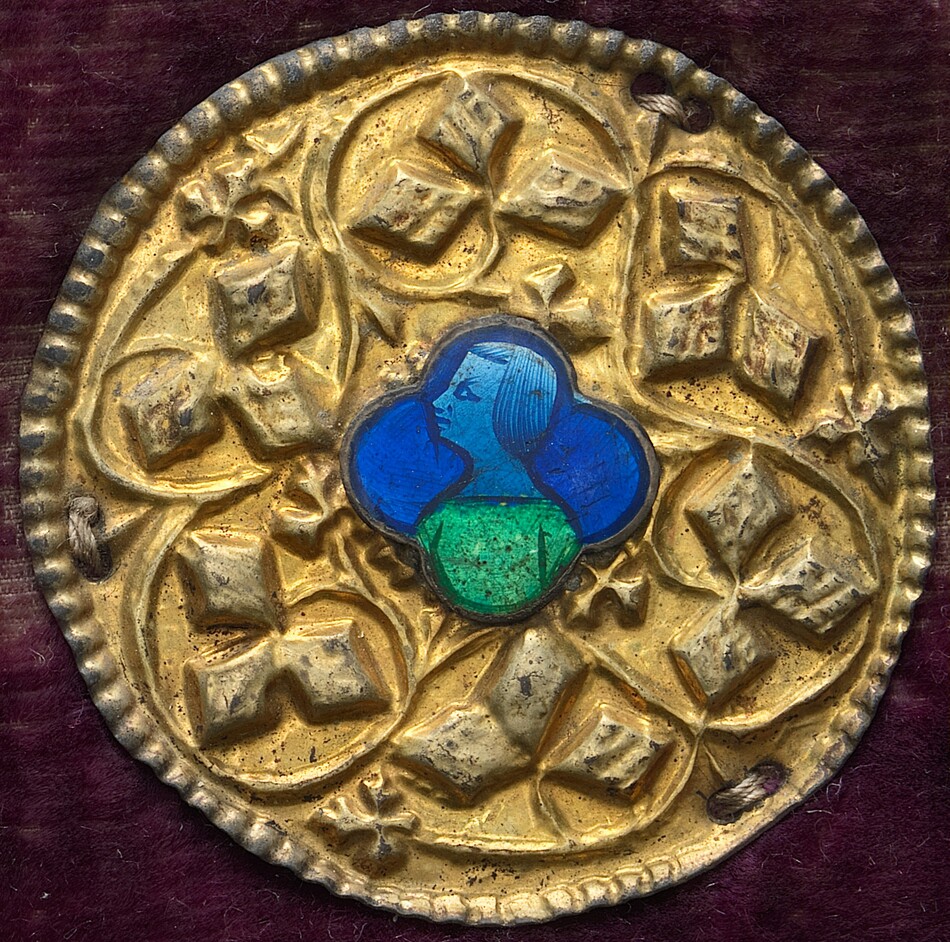 This is a photograph of a circular gilded object with an enamel centerpiece. The object is a richly decorated, golden disc featuring a central motif of enamel depicting a figure in profile with blue hair and a green tunic against a blue background. Surrounding the centerpiece is a symmetrical design of raised, geometric shapes. The edge of the object features a raised, beaded border, and the piece is set against a dark, warm-toned background, possibly velvet.