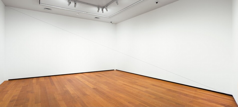 A taut, thin black cord stretches from the upper left to the lower right corner of a room white walls and hardwood floors. The cord is barely visible.