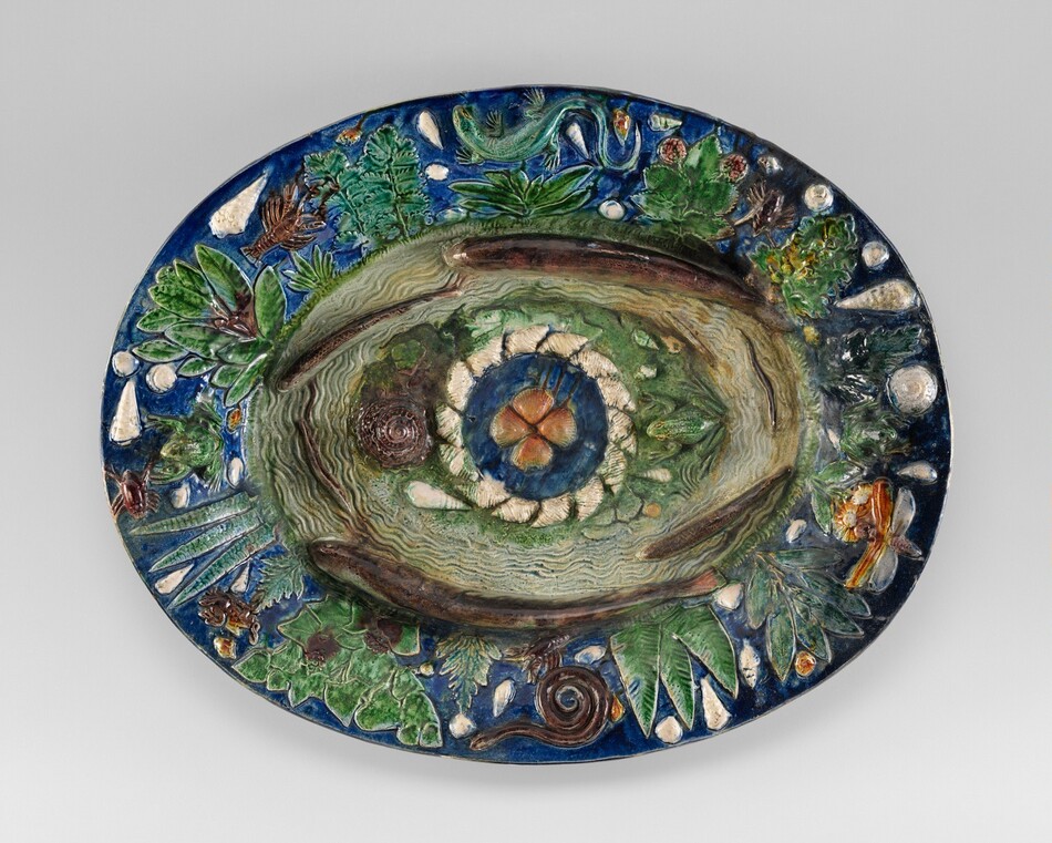 This is a photograph of an ornate ceramic platter. The surface of the platter is detailed with natural motifs, such as leaves, fruits, and small creatures like a snail and insects. In the center, there is a concentric circular design, surrounded by textures resembling wood and stone. The dominant colors are earthy greens and browns, with touches of blues, reds, and whites. The overall design captures the intricacies of nature in a three-dimensional form, showcasing pottery and ceramic glazing techniques.