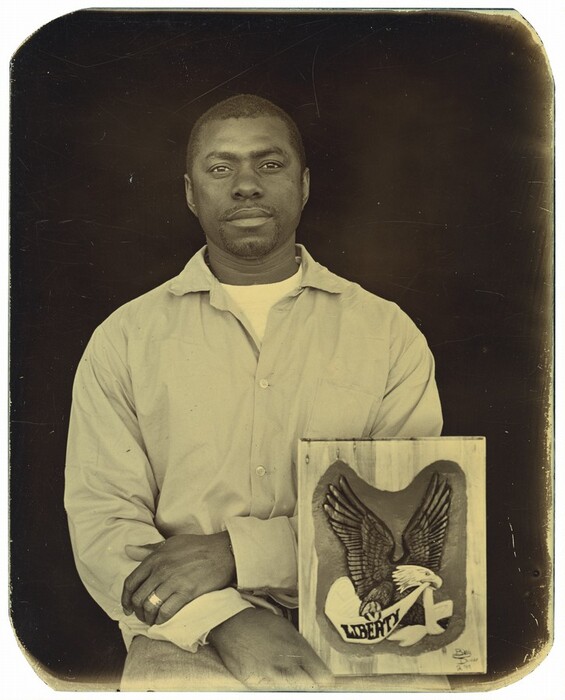 The photograph shows a man seated and facing forward. He has a calm expression on his face with short hair and a goatee. He is wearing a light-colored button-up shirt over a T-shirt, and his hands are resting on his lap. A wooden plaque next to him displays an eagle and the word "LIBERTY." The background is plain and dark.