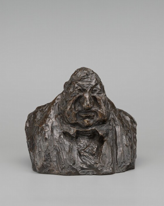 The sculpture depicts a human figure with a large head and exaggerated facial features, carved in a rough, textured style. The figure appears to be emerging from a rock-like formation, with the face being the most detailed part of the sculpture. The material of the sculpture is dark, likely bronze, with a metallic sheen and dark brown color. The surface is uneven and rugged, enhancing its substantial presence.