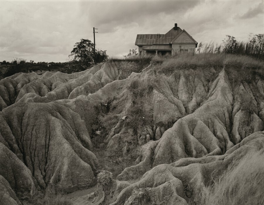 The image shows eroded terrain with rolling mounds in black and white. Closest to the viewer, the ground is rugged with distinct crevices. In the distance, there is an old house on the edge of the eroded landscape, silhouetted against a slightly overcast sky. The horizon aligns with the roof of the house, creating a flat line across the scene. The land appears naturally carved by erosion, giving it an organic and rugged appearance.