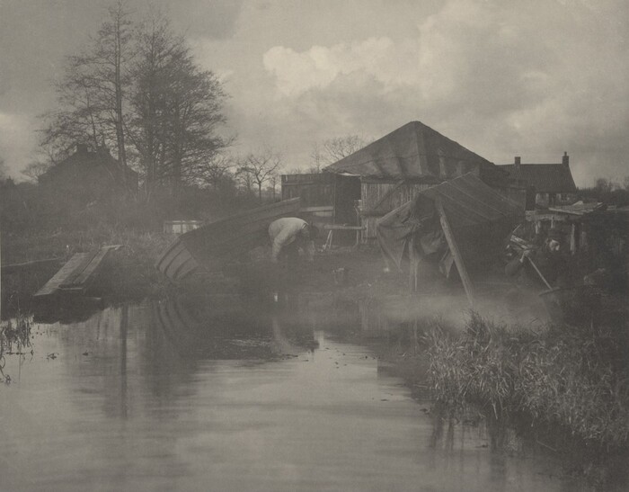 The image shows a calm waterway with reflections on the surface. Near the shore, a person is attending to an overturned boat. In the background, there are trees, buildings, and a dark-roofed structure. The scene is in black and white, with trees on the left and clouds in the sky. The atmosphere is quiet and slightly misty.