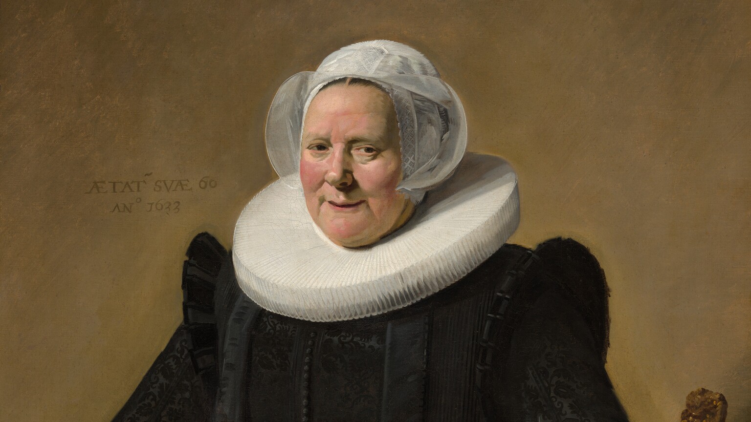 This vertical portrait painting shows a pale-skinned older woman wearing a full, long-sleeved black dress with a wide white ruff at her neck and white lace cuffs at her wrists. She sits in a curving, low-backed wood chair against a pale brown background. Her body and the chair are angled slightly to our left, and she looks directly at us. She has small eyes, pink, rounded cheeks, and a wide chin. Her lips are parted in a slight smile, and her hair is covered by a starched, sheer white cap that flares at the sides. The ruff around her neck is gathered in accordion-like, narrow figure-eight folds, and it extends flat and stiff, nearly to her shoulders. Close inspection reveals that the dress is woven with a black-on-black brocade pattern and has a line of small black buttons down the front. She holds a small brown leather book tooled with gold ornament in her right hand, on our left, and her other hand rests on the arm of the chair. She wears a gold ring on each hand. An inscription is painted to our left of her head: “AETAT SVAE 60 ANo 1633.”