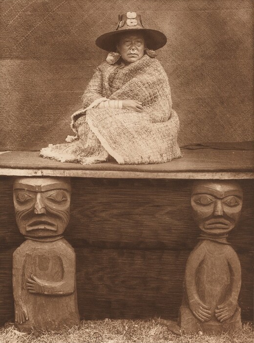 The image shows a person sitting cross-legged on a platform, covered in a woven blanket. The individual has arms resting on their knees, with prominent cheeks and a round nose ornament. They wear a wide-brimmed hat with round decorations, and bangles on their wrists. The platform has two carved wooden figures under the edge. The background is a textured surface.