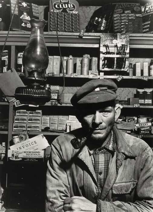 The image depicts a man from the waist up, leaning forward with both arms on a counter. He wears a cap tilted to one side, has a rough, unshaven face with pronounced facial features, and has short hair covered mostly by the cap. He wears a plaid shirt and a slightly worn coat or jacket. He holds a pipe in his mouth. The background is cluttered with various products on shelves, such as boxes, bottles, and tins with visible pricing. It appears to be an old shop or general store, with a vintage and utilitarian feel enhanced by invoices and product advertisements visible in the background.