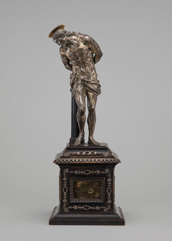 A bearded man wearing only a cloth tied around his hips bends toward a short column with his hands behind his back in this freestanding, silver sculpture. In this photograph, the man’s muscular body faces us, but his head turns sharply to the left as he looks down. A flat, starburst-like gold halo sits atop his long hair, which is pulled back to the nape of his neck. The black column is to our left, and it disappears behind the man’s hip. The tall pedestal has molding and decorative ornaments around a window on the front face. Material inside looks like tiny, fine chain links or a similar substance. The silver of the man’s body gleams with tones of muted pink and pale, slate blue where the light falls, especially across his chest and hips. The sculpture is photographed in front of a pale gray background.