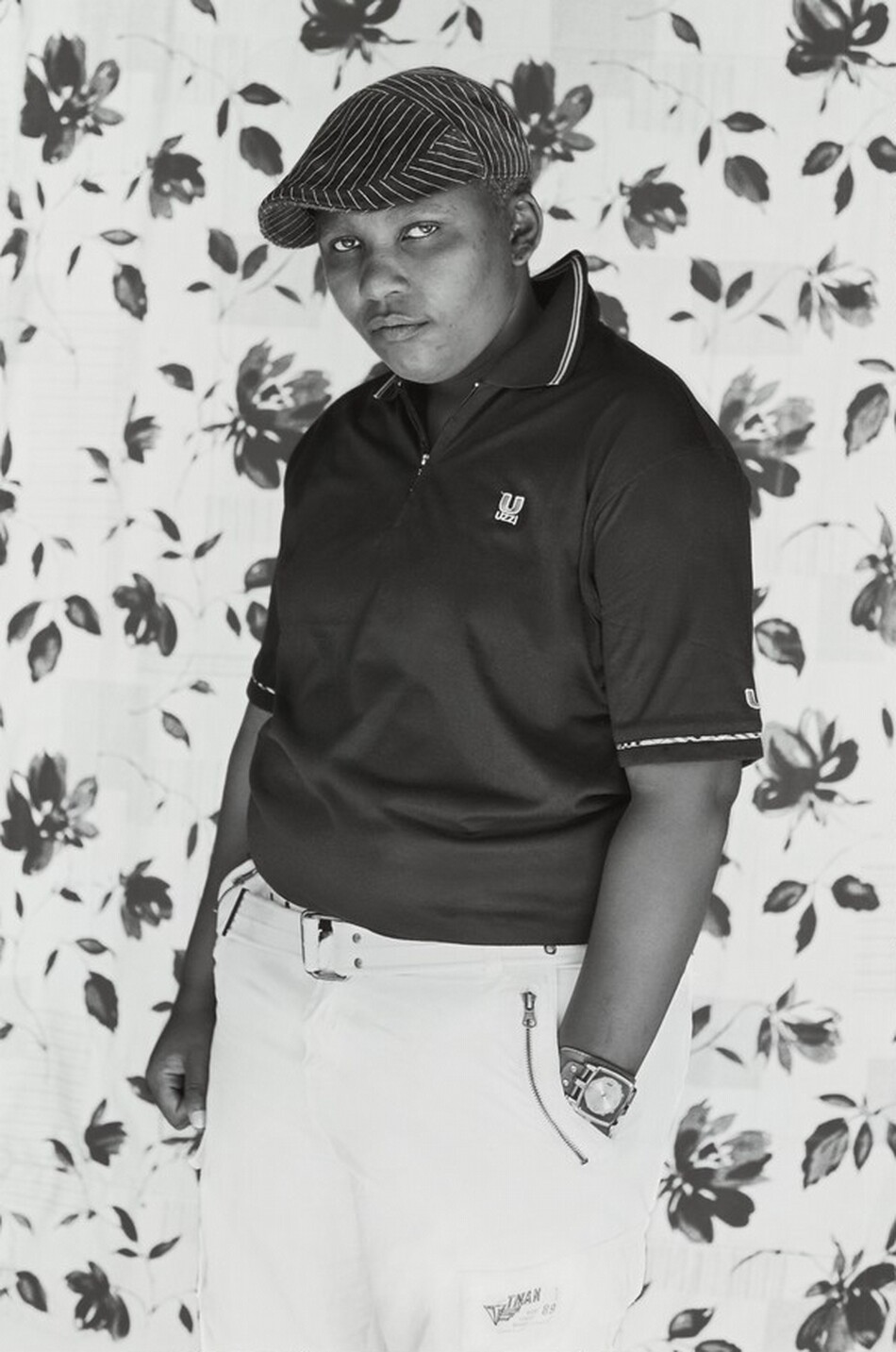 The image shows a person standing from the upper thigh upwards, slightly angled to the right. They are wearing a dark polo shirt with a collar and a small emblem on the left side of their chest. Their head is slightly tilted forward, and they have a short haircut styled with a striped flat cap. Their gaze is directed towards the camera with a neutral expression. They are also wearing light-colored trousers, with a visible belt and a watch on their left wrist. The background consists of a patterned floral curtain with large flowers and leaves in contrasting tones.