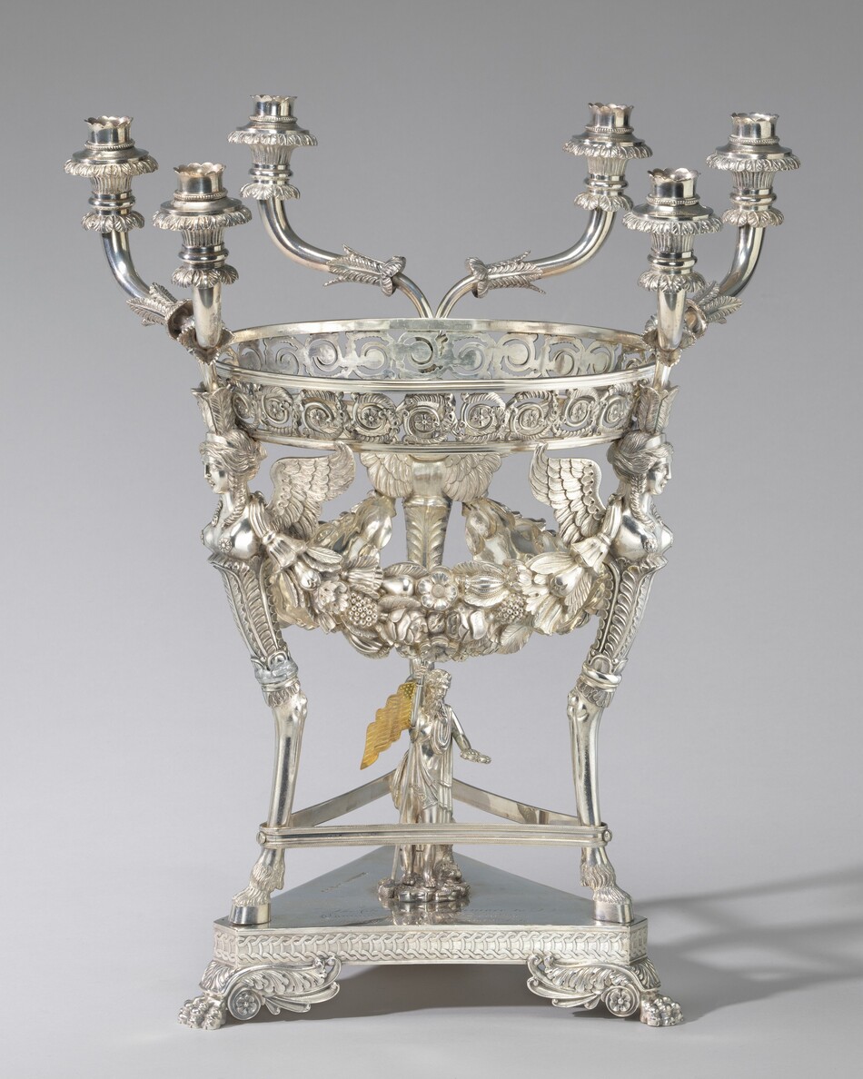 The sculpture is a candelabrum with multiple intricate branches for holding candles. It features a large circular bowl with floral patterns, garlands, and scrollwork. Winged female figures support the bowl, adding an ornate touch. The base incorporates decorative elements and rests on legs resembling lion paws. The sculpture is made of silver, giving it a polished appearance. It showcases exceptional craftsmanship and decorative motifs, exuding elegance. The design suggests it is intended as a centerpiece.