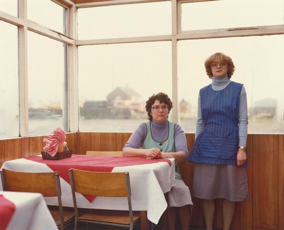 The image depicts two women indoors. One woman is seated at a table, wearing a light green apron and a light purple turtleneck sweater, with short, curly brown hair and glasses. The other woman is standing to the right, with shoulder-length wavy blonde hair, glasses, and wearing a blue striped apron over a light purple turtleneck sweater and gray skirt, along with a wristwatch on her left wrist. The setting includes wood-paneled walls, large windows showing a blurred outdoor scene with a house and parking area, and a table with a white and red tablecloth holding a basket with napkins and condiment bottles.