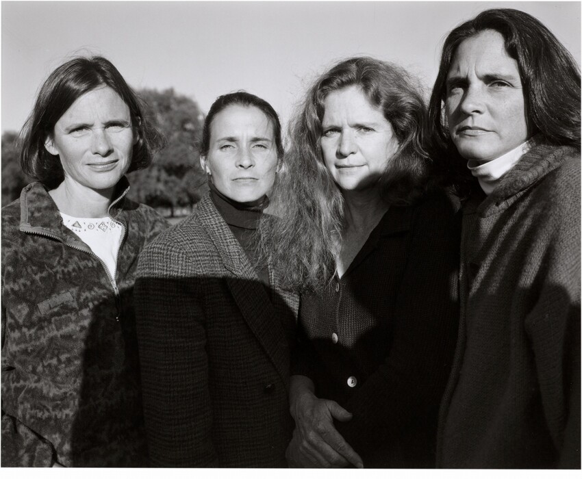 Four women are depicted from the waist up, standing closely together in a line facing the camera. Each woman has distinct facial features. The woman on the left has short, dark hair parted to one side, the second woman has pulled-back hair, the third woman has long, curly hair, and the woman on the right has long, straight dark hair. They are dressed in layered clothing appropriate for cooler weather, including sweaters and coats. One woman has a patterned jacket, and another wears a turtleneck. They have neutral expressions. The background shows a clear sky, some trees, and sunlight casting a slight shadow on two of the women.