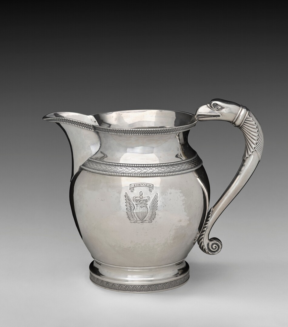 The image shows a large, polished silver pitcher with intricate bead detailing around its neck and base. The handle is sculpted in the form of a stylized animal, likely an eagle, with its head at the top of the handle and its body merging into the base. The color palette is predominantly silver, with various shades created by reflections and shadows, giving it a metallic shine. In the center of the pitcher, there is an engraved emblem or crest positioned prominently on the front face. The spout of the pitcher extends slightly forward, leading the eye from left to right across the image, while the handle curves from the back towards the right side.