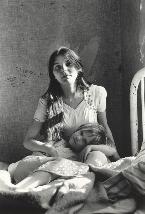 The image shows two individuals: a woman and a child seated. The woman sits on a bed with the child's head resting on her lap. The woman has long, dark hair and a calm expression, wearing a light-colored dress with floral embroidery. The child, facing the camera, has shorter, light-colored hair and looks relaxed in a light top and covered by a blanket. In the background is a plain wall with the words "SHARON T." and "Bob" written on it, and a bedpost is visible to the right. Light appears to be coming from the right side, casting shadows on the wall and bed, creating a simple and intimate atmosphere.