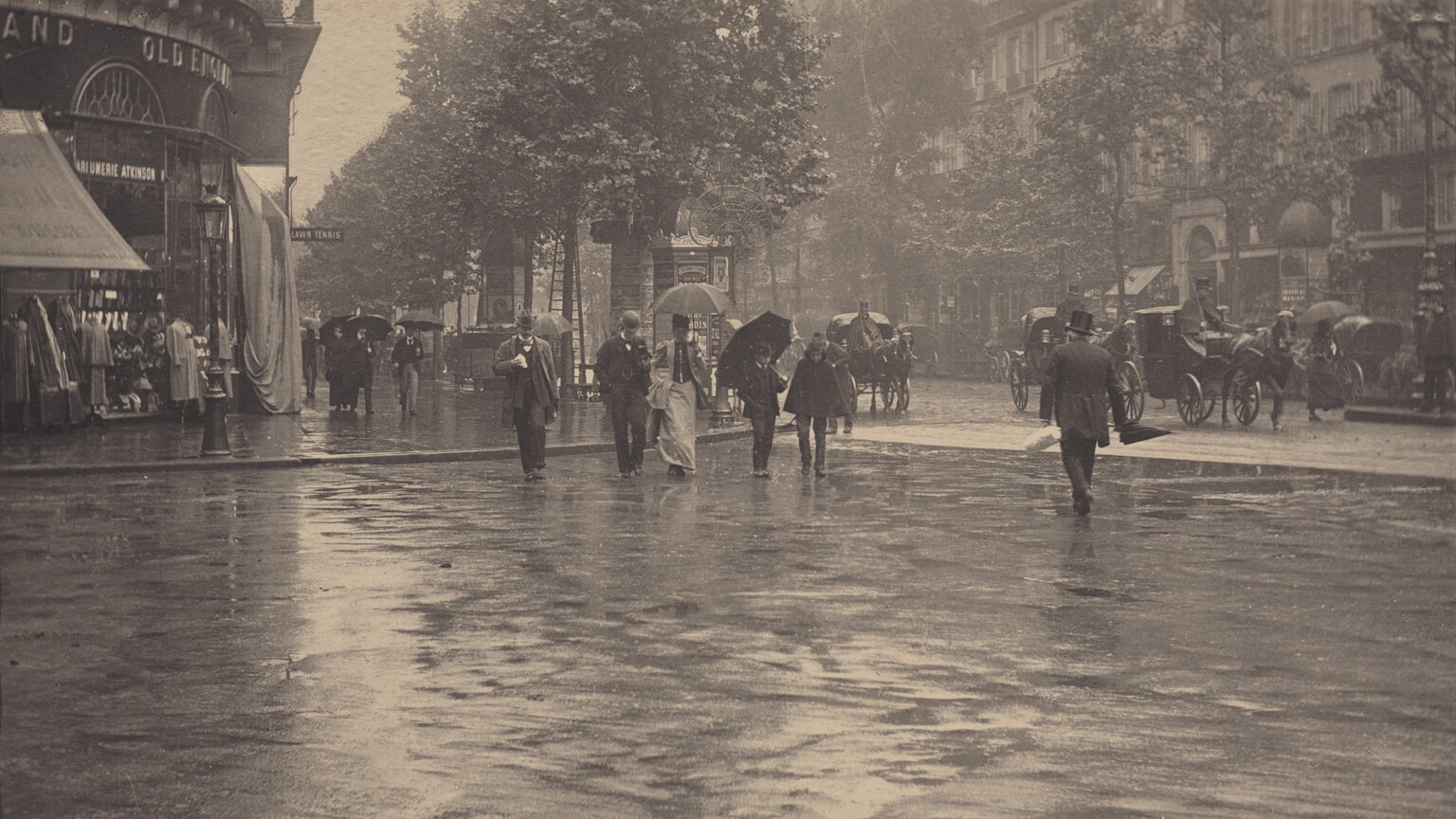 In this black and white horizontal photograph, people and horse-drawn carriages travel along and cross a rain-slicked, urban street. The photograph is roughly divided in half horizontally so the lower half shows a the wet pavement of a wide intersection, momentarily clear of people, where two streets meet in a T intersection. The upper half shows shops and buildings along a tree-lined street at the corner to our left, and along the long, cross street to our right. On the corner, clothing and other wares are displayed outside under an awning , while another display is covered over with a tarp. There are about ten people are on foot crossing the street toward us, while several horse-drawn carriages with large, spoked wheels, driven by caped and top-hatted men, pass through or are parked alongside the curb on the far side of the street. Some of the pedestrians hold up dark umbrellas, while only one man, without an umbrella, crosses the street walking away from us. All of the men wear dark suits and hats. The one woman crossing with the group wears a long skirt and matching thigh-length coat over a contrasting buttoned bodice. She holds up her skirts in one hand and her umbrella in the other. At the far corner, several tall multi-paneled kiosks with domed tops are papered with posters. Across the street is an uninterrupted row of buildings and shops with rows of shuttered windows above. A small triangular slice of sky can be seen between the building at the left and the trees lining the street; its reflection is mirrored on the wet street.
