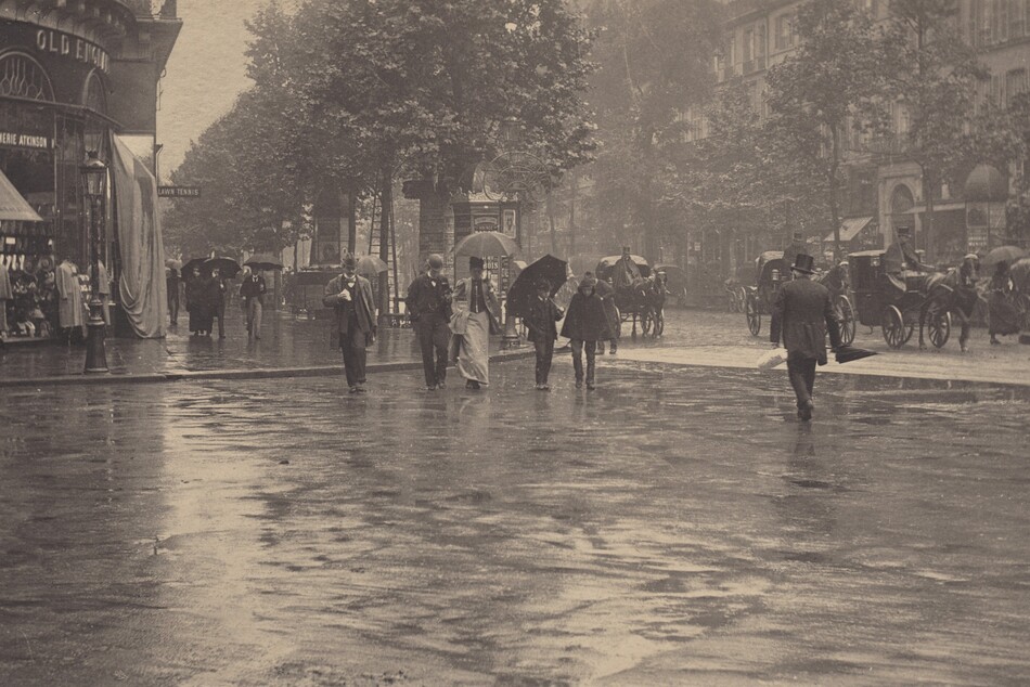 In this black and white horizontal photograph, people and horse-drawn carriages travel along and cross a rain-slicked, urban street. The photograph is roughly divided in half horizontally so the lower half shows a the wet pavement of a wide intersection, momentarily clear of people, where two streets meet in a T intersection. The upper half shows shops and buildings along a tree-lined street at the corner to our left, and along the long, cross street to our right. On the corner, clothing and other wares are displayed outside under an awning , while another display is covered over with a tarp. There are about ten people are on foot crossing the street toward us, while several horse-drawn carriages with large, spoked wheels, driven by caped and top-hatted men, pass through or are parked alongside the curb on the far side of the street. Some of the pedestrians hold up dark umbrellas, while only one man, without an umbrella, crosses the street walking away from us. All of the men wear dark suits and hats. The one woman crossing with the group wears a long skirt and matching thigh-length coat over a contrasting buttoned bodice. She holds up her skirts in one hand and her umbrella in the other. At the far corner, several tall multi-paneled kiosks with domed tops are papered with posters. Across the street is an uninterrupted row of buildings and shops with rows of shuttered windows above. A small triangular slice of sky can be seen between the building at the left and the trees lining the street; its reflection is mirrored on the wet street.