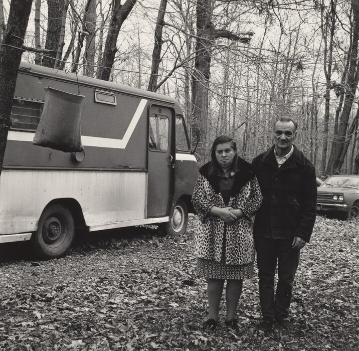 The image shows two people standing side by side, captured from the knees up. The woman on the left has medium-length straight hair and relaxed facial features. She is wearing a patterned overcoat, a dress, and dark shoes. The man on the right has short hair combed back and a mustache, and he is wearing a dark-colored coat, a collared shirt, and trousers. They are in a wooded area with leafless trees and fallen leaves on the ground. There is a vintage styled van with a bold diagonal stripe to the left of them, and a car partially visible among the trees. A hanging object is suspended from a tree branch to the left of the van.