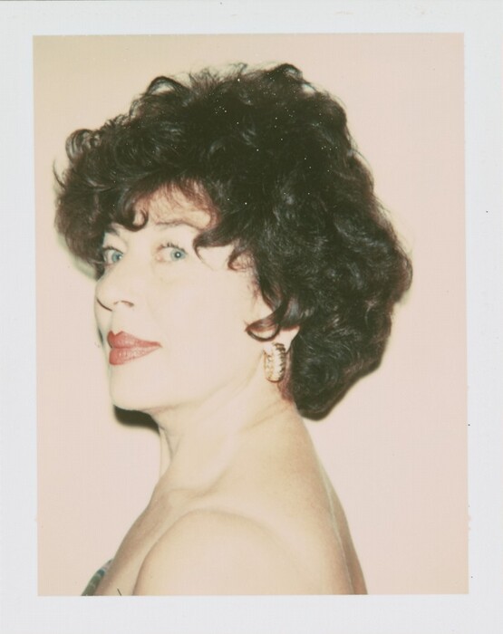 The image depicts an older woman with her upper torso visible. She is facing the left side of the image, emphasizing her profile. The woman has blue eyes, a defined nose, and red lipstick. Her dark, curly hair is styled voluminously, and she wears large gold hoop earrings. She is dressed in a strapless top with one shoulder exposed. The background is a light-colored wall.
