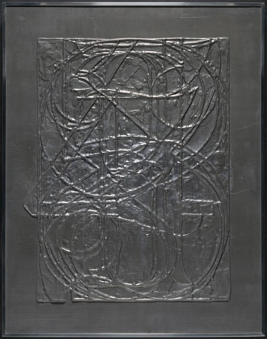 The sculpture is an abstract relief design made of metallic material. It features a network of interweaving and overlapping lines and curves, creating a complex web or layered grid. The surface is a dark, matte silver color with raised lines forming a textured pattern that adds depth. Light plays across the sculpture, highlighting its form and texture. The backdrop is smooth in contrast to the intricate and raised lines of the sculpture.