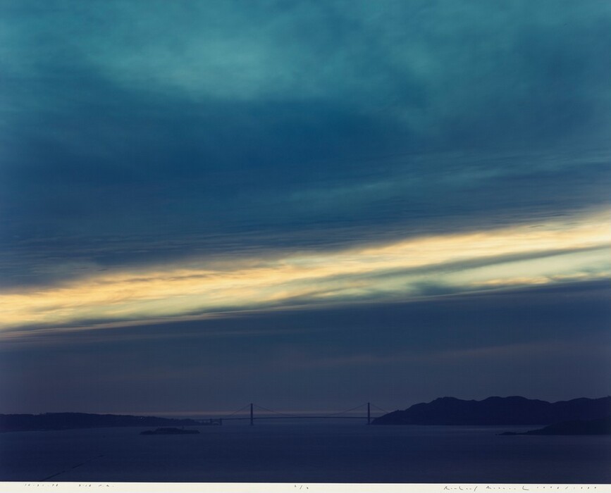 The image features a wide-angle view of a landscape where water meets the sky. The horizon is low in the composition, with a bridge visible in the distance. Landforms in the background are dark and minimally detailed, contrasting with the lighter water and sky. The color palette includes deep blues and soft yellows, with gradations in the sky from dark to light. Clouds are depicted with diffuse brushstrokes, creating a soft effect. Light and shadow play across the sky and landscape.