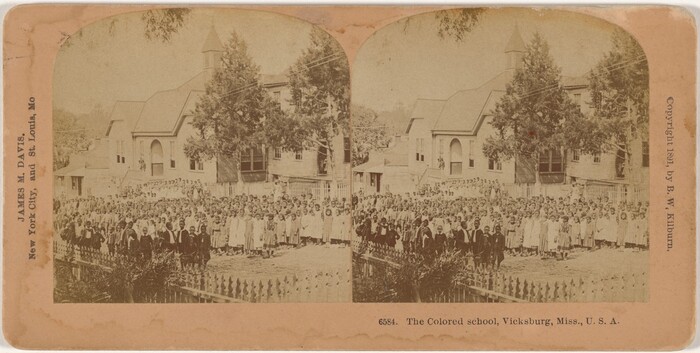 This is a photograph of a group of children standing in front of a wooden church building. The children are dressed in a mix of light and dark colored clothing. The background includes the church, trees, and a white picket fence.