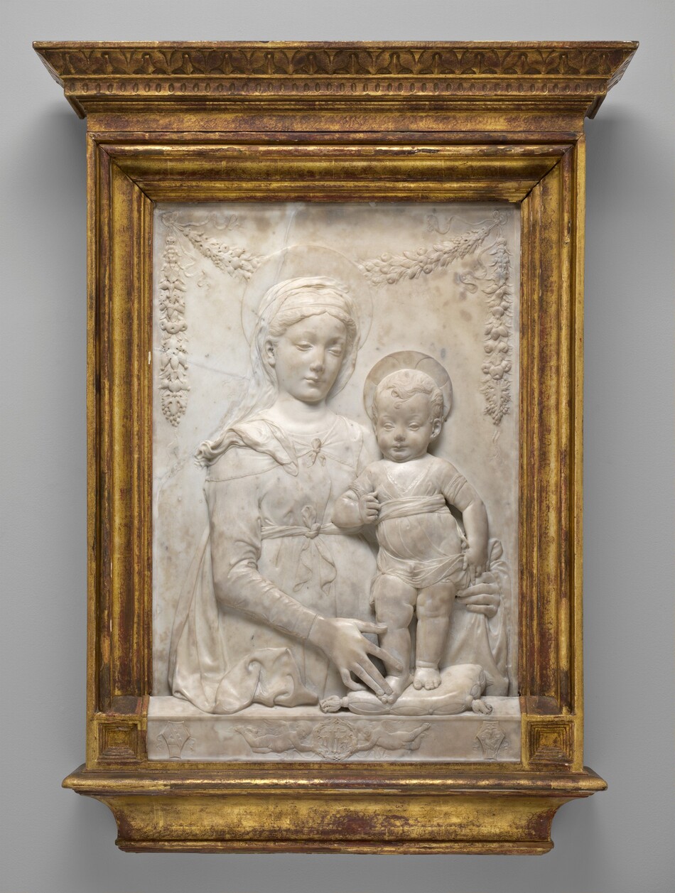 The sculpture depicts a relief of a mother holding a small child. Both figures have halos. The sculpture is made from a light-colored material and has delicately carved details. The figures are framed by a rectangular, ornate frame with a gilded, golden finish. The mother figure has serene features. She supports the child with one hand while the child stands confidently. This sculpture is mounted on a wall.