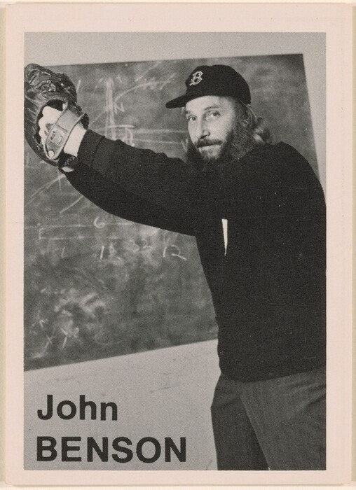 The image depicts a man standing with his arms raised, holding a baseball glove in his left hand. He has a full beard, long hair, and a dark baseball cap with a visible "B" emblem. He wears a dark long-sleeved shirt, and the background shows a chalkboard with various markings and the words "John BENSON" at the bottom.
