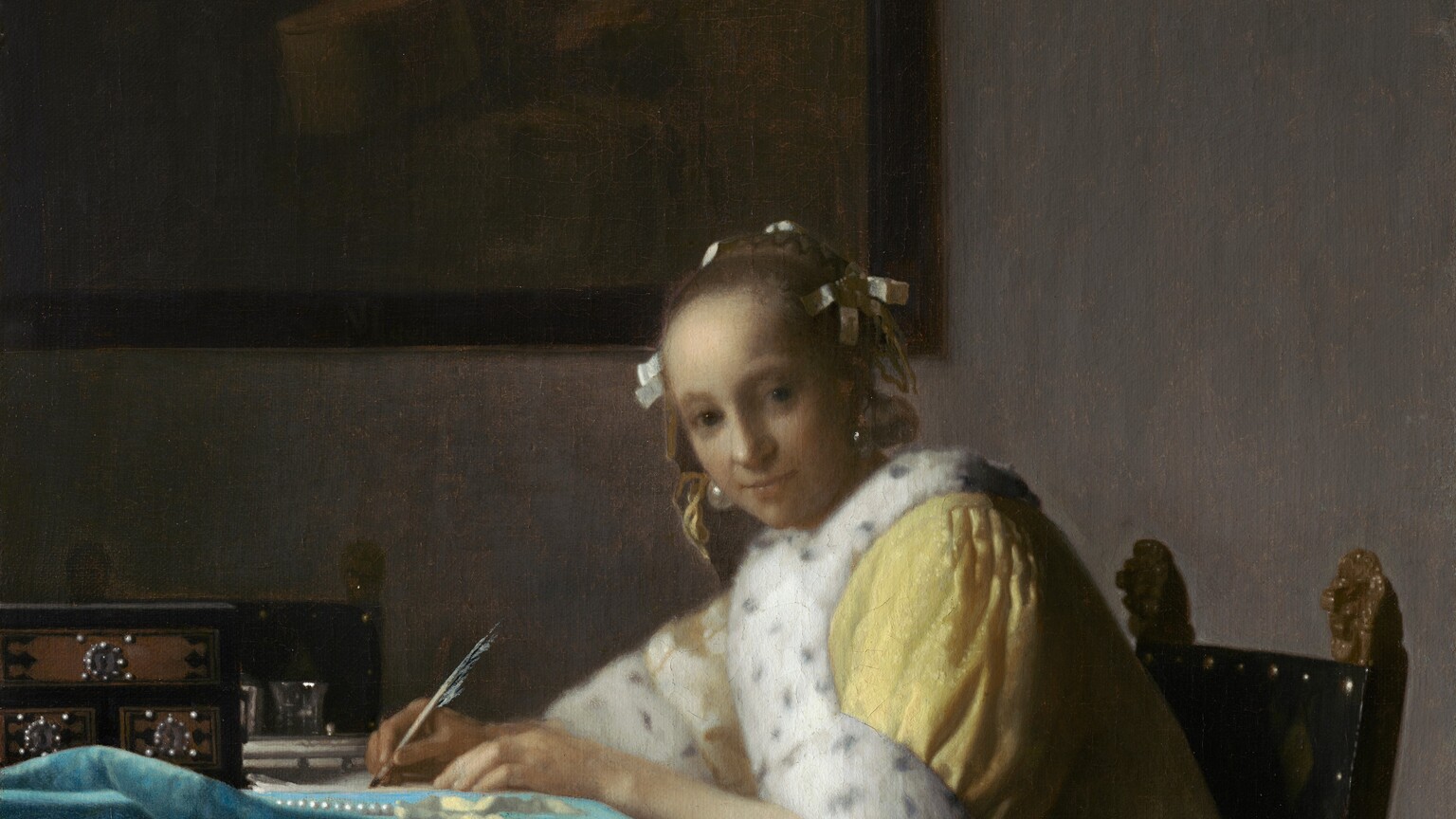 Shown from about the knees up, a pale, smooth-skinned woman in a fur-lined yellow jacket looks out at us as she sits writing at a table in this vertical painting. The woman’s body faces the table to our left. She turns her head to gaze at us from the corners of her dark gray eyes under faint brows. She has a wide nose, and her pale lips are closed. Her light brown hair is pulled back and held in place with white bows, and gleaming teardrop-shaped pearl earrings dangle from her ears. Her lemon-yellow jacket is trimmed with ermine fur, which is white with black speckles, at the cuffs and down the front opening. A full, elephant-gray skirt falls to the floor beneath the jacket. Both hands rest on the table, and she holds a quill in her right hand, farther from us, on a piece of paper. She leans forward in her wooden chair. The back panel of the chair is covered in black fabric and lined with brass studs. Two gilded finials, carved into lions’ heads, face the woman’s back with mouths open. The table is covered with a celestial-blue cloth crumpled near the left edge of the canvas. On the table are a strand of pearls, a pale yellow ribbon, and a black box with three brown panels studded with pearls around silver keyholes. Two pewter gray vessels are visible just beyond it, in front of a second chair, which faces us. On the putty-gray wall behind the woman, a framed painting hangs in the upper left quadrant of the composition. The painting-within-the painting is done in muted tones of brown and shows a cello and other unidentifiable objects.