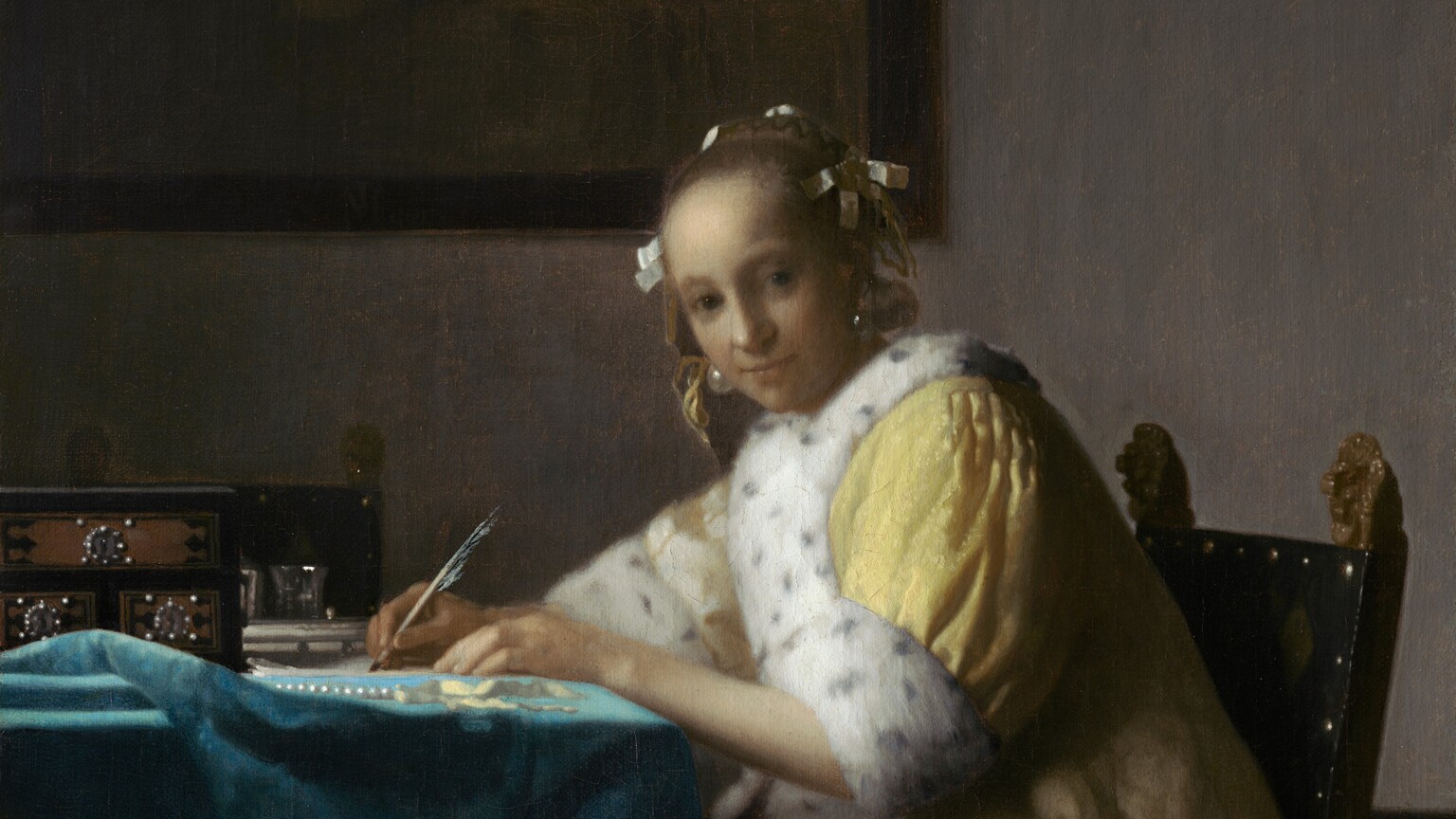 Shown from about the knees up, a pale, smooth-skinned woman in a fur-lined yellow jacket looks out at us as she sits writing at a table in this vertical painting. The woman’s body faces the table to our left. She turns her head to gaze at us from the corners of her dark gray eyes under faint brows. She has a wide nose, and her pale lips are closed. Her light brown hair is pulled back and held in place with white bows, and gleaming teardrop-shaped pearl earrings dangle from her ears. Her lemon-yellow jacket is trimmed with ermine fur, which is white with black speckles, at the cuffs and down the front opening. A full, elephant-gray skirt falls to the floor beneath the jacket. Both hands rest on the table, and she holds a quill in her right hand, farther from us, on a piece of paper. She leans forward in her wooden chair. The back panel of the chair is covered in black fabric and lined with brass studs. Two gilded finials, carved into lions’ heads, face the woman’s back with mouths open. The table is covered with a celestial-blue cloth crumpled near the left edge of the canvas. On the table are a strand of pearls, a pale yellow ribbon, and a black box with three brown panels studded with pearls around silver keyholes. Two pewter gray vessels are visible just beyond it, in front of a second chair, which faces us. On the putty-gray wall behind the woman, a framed painting hangs in the upper left quadrant of the composition. The painting-within-the painting is done in muted tones of brown and shows a cello and other unidentifiable objects.