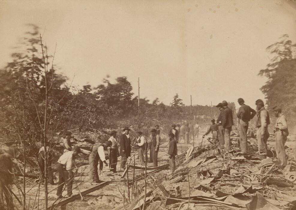 This is a photograph of several people working on a railway track in a wooded area. The scene shows individuals engaged in laborious activities, surrounded by trees and bushes. The ground is littered with debris and metal parts, suggesting construction work. Various tools are visible, and some individuals seem to be supervising or assisting with the task. The image captures a moment of manual labor in a rugged environment.