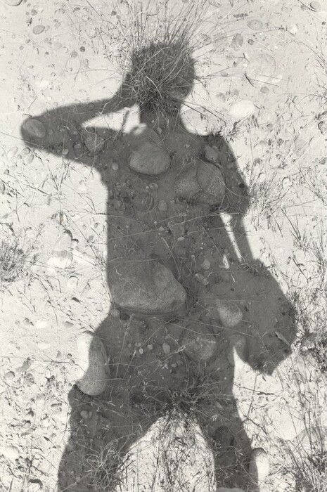 The image shows a shadowy silhouette on a textured surface. The silhouette has distinct lines and curves, resembling a human-like form. The abstract representation includes a large shadow that looks like a person holding something up to their face. The shadow is cast on a ground with gravel, stones, and sparse patches of grass.