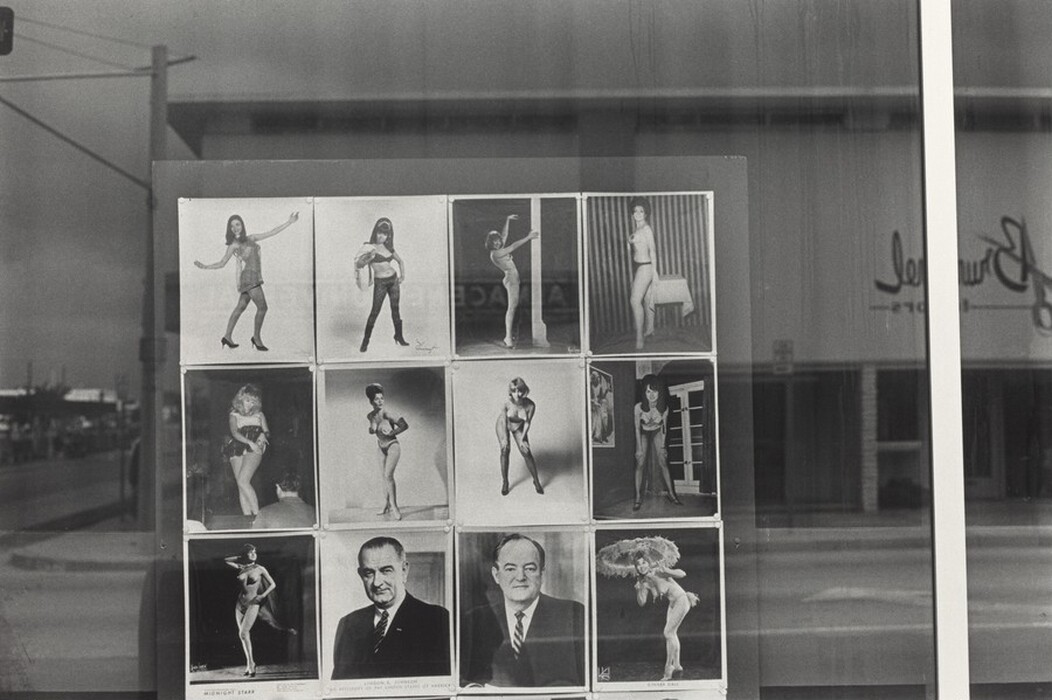 This is a photograph of a window display featuring twelve photographs arranged in a grid format. The images predominantly showcase women in different poses and attire, with two photographs also depicting men in formal attire. The window reflects an urban environment outside, including a street and architectural elements, as well as cursive signage. The black-and-white medium highlights details in the images and reflections.