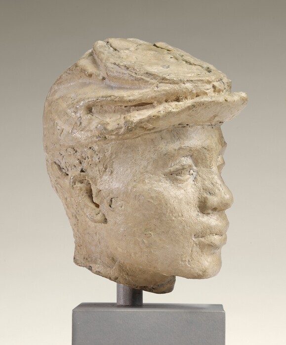 This sculpture depicts a human head of a young person wearing a hat. The facial features are detailed, including the nose, mouth, and eyes. The sculpture is made from a light-colored material that appears smooth yet slightly textured. The hat's outline and folds are visible, and the head is mounted on a small pedestal. The piece is portrayed in a three-dimensional format, providing a lifelike representation.