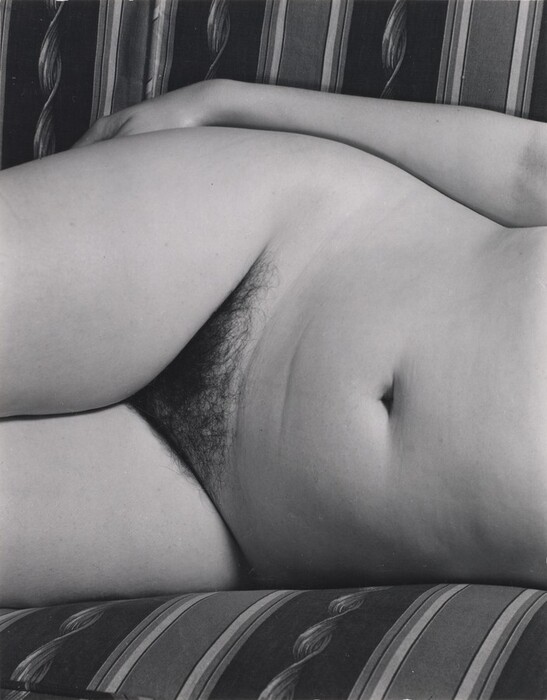 The image shows a close-up section of a human body from the shoulders to the upper thighs, focusing on the torso. The person is lying down, partially on their side, on a fabric surface with a striped pattern. The fabric has vertical stripes in varying widths and shades. The image does not include the face. There is no clothing visible in this section of the body. The setting appears to be indoors, suggested by the cushioned fabric surface on which the person rests. There are no visible objects or jewelry associated with the person in this image.