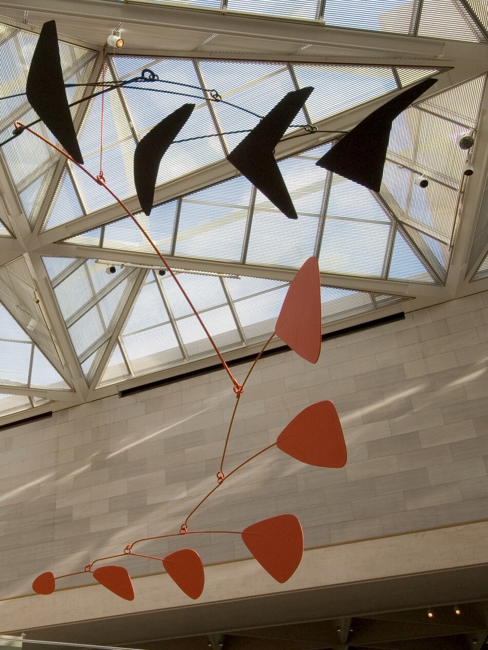 Six black, wedgelike shapes, one dark blue arrowhead shape, and six red triangular shapes with rounded corners are connected with thin rods to form this abstract sculpture. In this photograph, we look up at the piece to where it hangs from the glass ceiling of a light-filled interior space with marble walls. The paddle-like shapes are attached to the end of the curved rods, which are linked together like branches. In our view, the black arm curves up across the top of the picture and the red arm curves down into the lower right corner, and to our left. Each red paddle is smaller as they move from the central armature to the end of the branch.