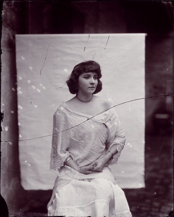 The image shows a woman seated from the waist up in a classic portrait style. She is facing slightly to the right with her hands crossed on her lap. Her facial features are clearly defined with dark eyes and calm expression. She has dark, wavy bob hair. She wears a light-colored off-the-shoulder garment with lace detailing, a pearl necklace, and earrings. The background is a plain white backdrop with cobblestone ground and shadowy areas in the upper corners.